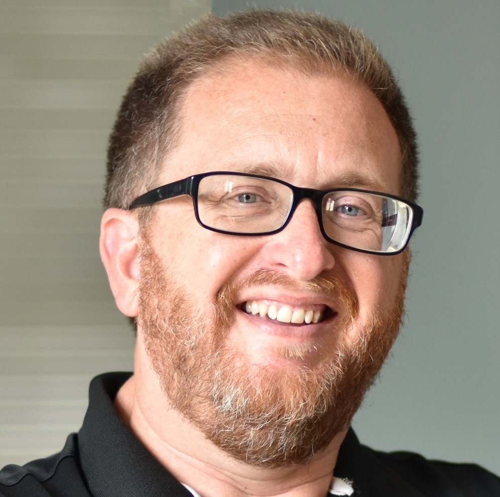 A bespectacled man with sandy reddish hair, mustache, and beard smiles for the camera.