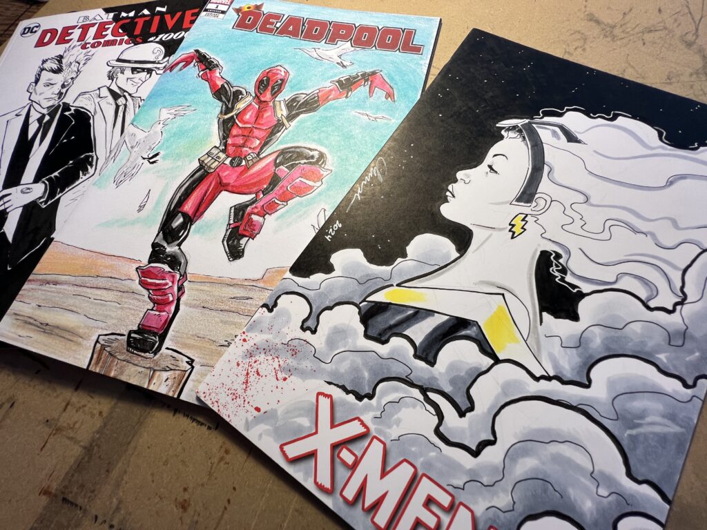 IMG_2712 Three comic book covers are spread out on a drawing table. One features a black and white drawing of Batman villains Two Face and the Riddler. The middle book has a full-color sketch of Deadpool in a Karate Kid-style crane kick pose. The third book is a profile view of X-Men character Storm, with clouds billowing up around her.