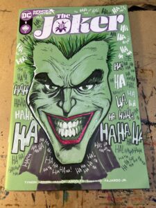 A green comic book sketch cover, with a hand-drawn portrait of a smiling Joker and the words "Ha ha ha" written chaotically across the background.