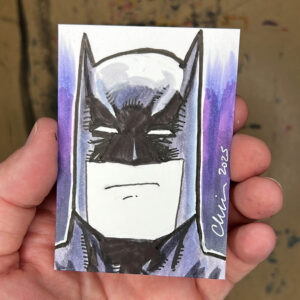 A hand is holding a sketch card with a drawing of the comic book character Batman on it.